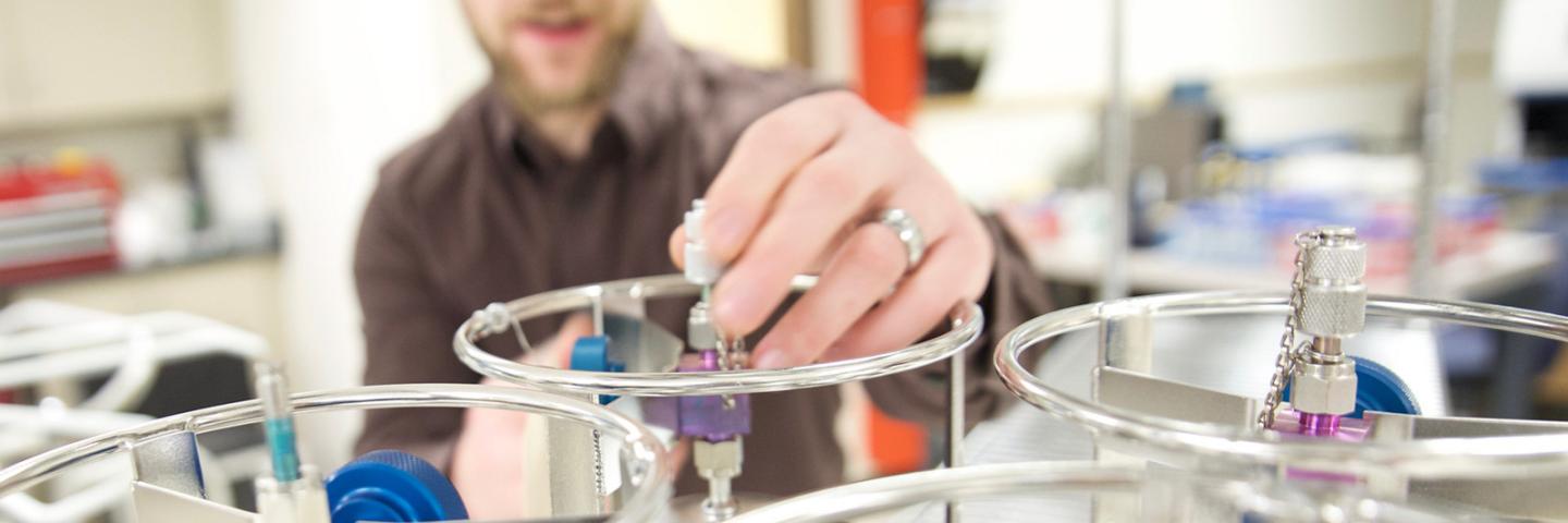 A man in a lab turns a knob on top of a metallic cannister.