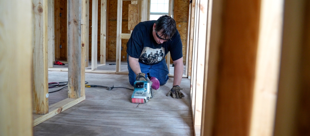 A man using a belt sander on the floor of a building being constructed.