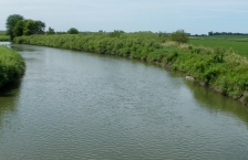 Wide, curving stream running through flat grasslands and fields. 