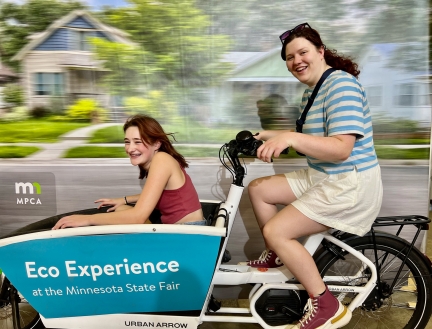 Girl sitting on electric bike with second girl in the front cargo box in front of a blurry photo background to look like bike is in motion.